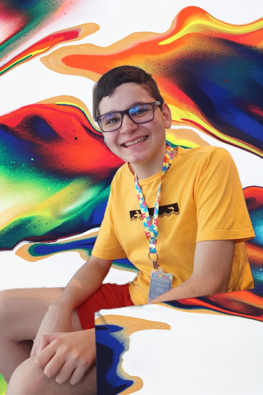 Kewen Lucas sorrindo, usando camiseta amarela e cordão colorido, diante de um fundo artístico colorido.